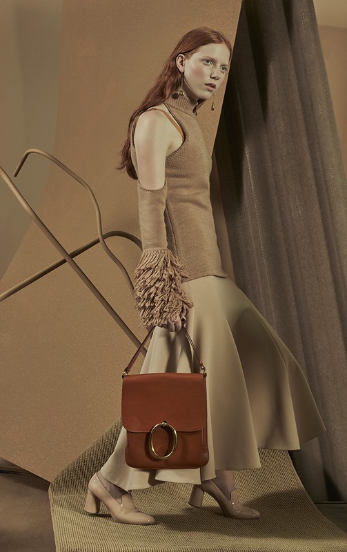 Fashion shot of a red-haired woman in a sleeveless knit top and long skirt, holding a purse with metallic gold hardware, indoors.