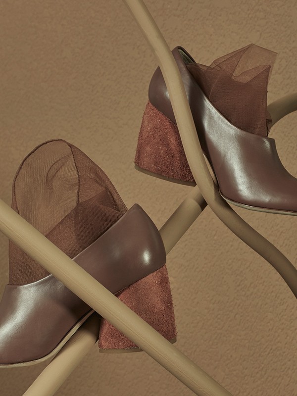 Fashion still life of a pair of heeled shoes with brown leather and suede details, draped on light wood sticks against a warm brown backdrop.