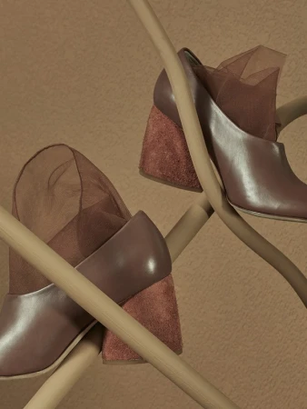 Fashion still life of a pair of heeled shoes with brown leather and suede details, draped on light wood sticks against a warm brown backdrop.
