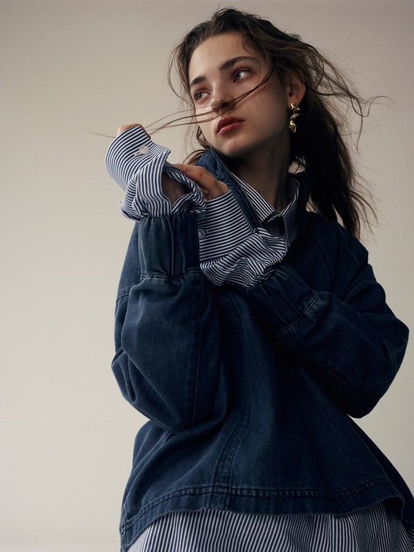A fashionable young woman with wavy brunette hair poses in a blue denim jacket and striped shirt against a neutral-toned backdrop.
