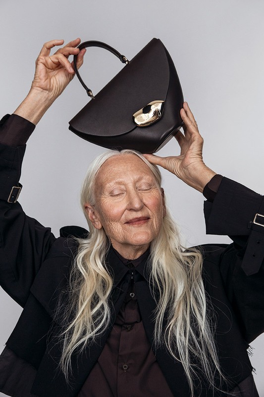 Older woman with white hair holds a dark brown leather handbag with gold hardware above her head in a studio fashion shot.