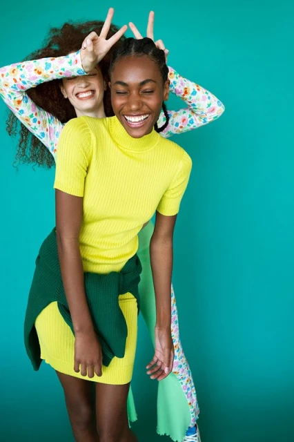 Two young women playfully pose against a teal backdrop, one with curly hair making peace signs, the other with braided hair smiling broadly.