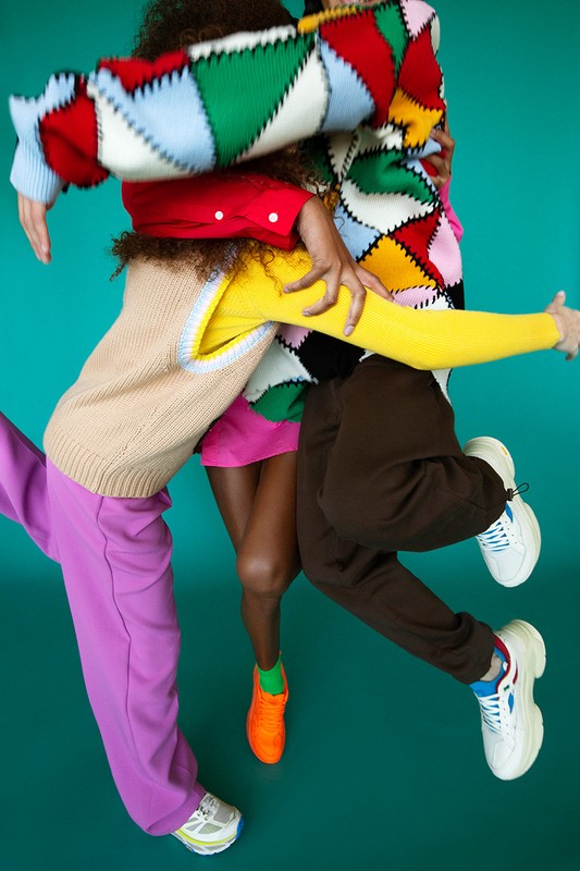 A group of people wearing colorful sweaters and pants, jumping in the air against a teal background in a fashion editorial photograph.