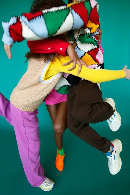 A group of people wearing colorful sweaters and pants, jumping in the air against a teal background in a fashion editorial photograph.