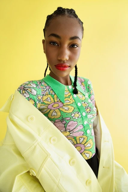 Portrait of a dark-skinned woman wearing a green and pink floral shirt, a yellow jacket, and red lipstick against a yellow background.