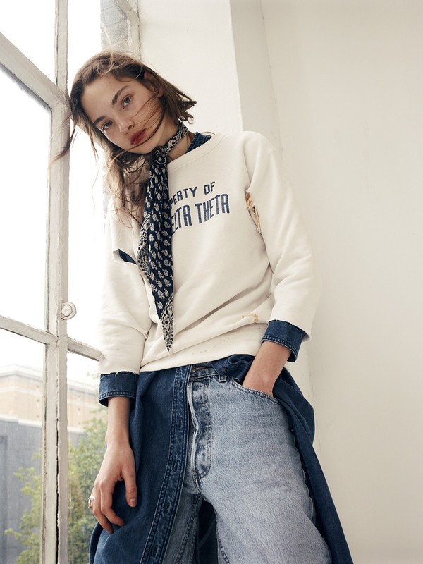 Young woman poses by a window wearing a cream sweatshirt, dark patterned scarf, and wide leg two-tone denim jeans in studio lighting.
