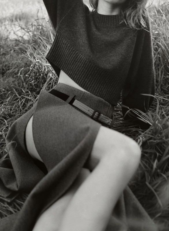 Black and white shot of a woman in a cropped sweater and skirt with a slit, posing in tall grass, soft lighting, medium shot.