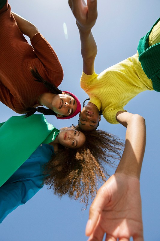 Low-angle shot of three young women looking down, wearing colorful tops and casual clothes against a bright blue sky on a sunny day.