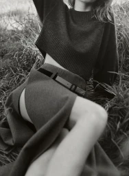 Close-up monochrome editorial fashion shot of a woman in a dark cropped knit sweater and high-waisted belted skirt reclining outdoors in tall grass