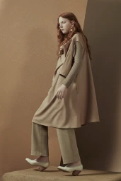 Red-haired female model wearing layered beige tunic and trousers posing against geometric textured studio backdrop.