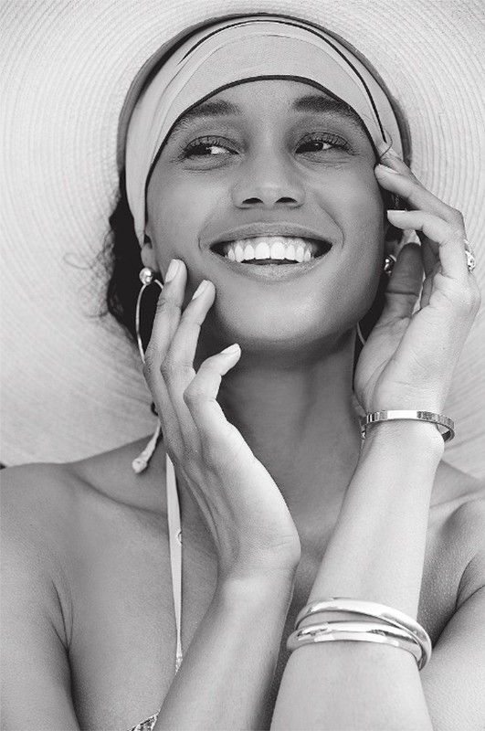 Black and white portrait of a smiling woman wearing a wide-brimmed hat and headscarf, with hands near her face and jewelry visible.