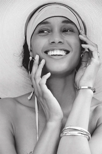 Black and white portrait of a smiling woman wearing a wide-brimmed hat and headscarf, with hands near her face and jewelry visible.