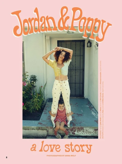 Woman standing behind a child sitting on a stoop in front of a door, wearing fashionable clothing in a pink-themed outdoor setting.