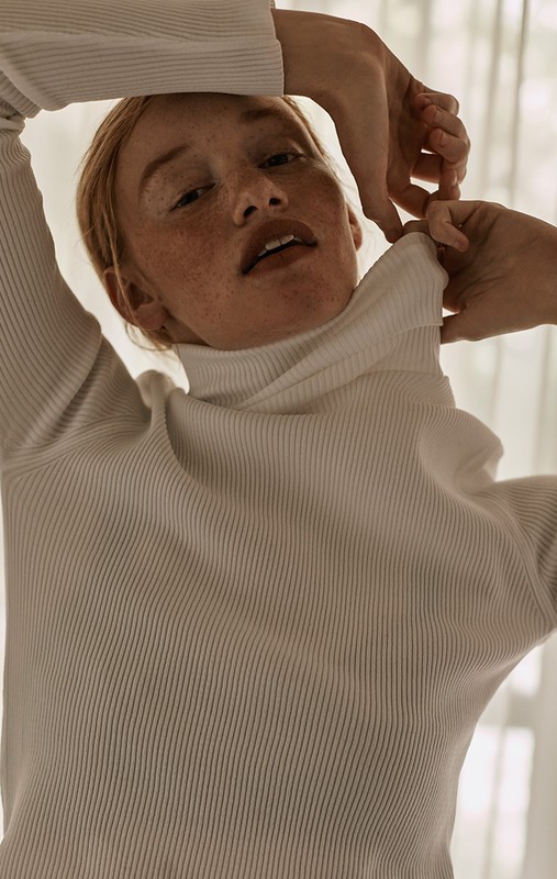 Redhead woman in white ribbed turtleneck sweater poses with hands framing face, soft light, and neutral background indoors.