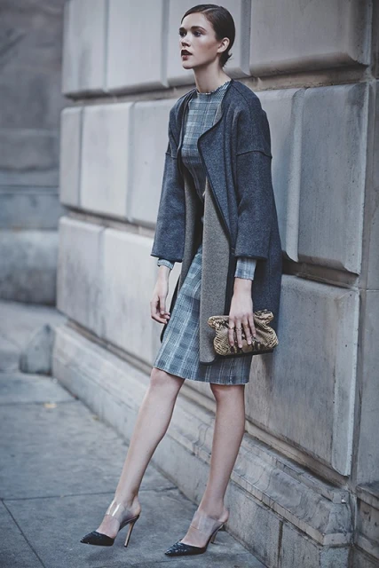 A young adult woman stands against a textured wall wearing a grey coat and plaid dress, holding a woven clutch and wearing transparent heels.