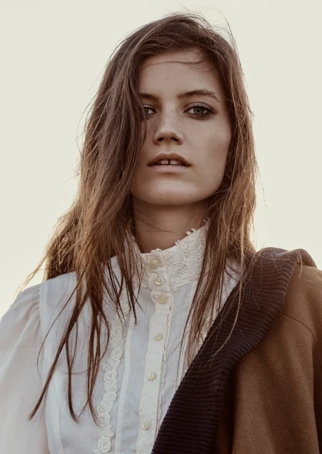 Close-up portrait of a young adult woman with long brown hair, wearing a white lace blouse and brown coat against a soft, diffused background.