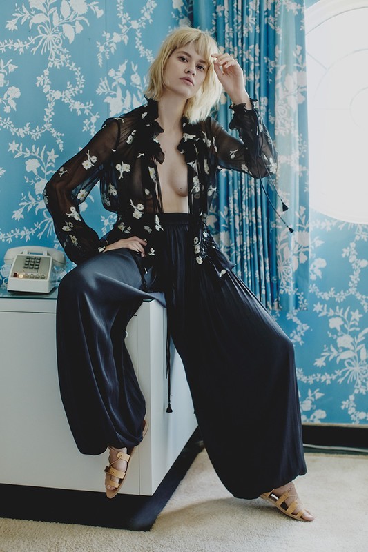 Young woman in fashionable black wide-leg pants and sheer floral top poses indoors with vintage phone on a table, against blue floral wallpaper.