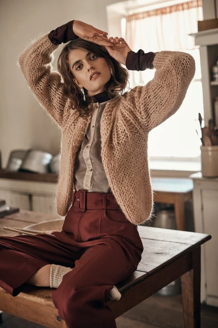 A young woman with curly brown hair sits on a wooden table in a kitchen, wearing a pink sweater, dark red pants, and neutral-toned shirt.