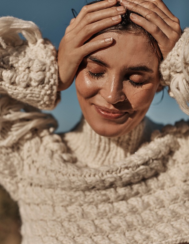 Close-up portrait of a smiling woman with her hands on her head, wearing a textured sweater against a blue sky backdrop.