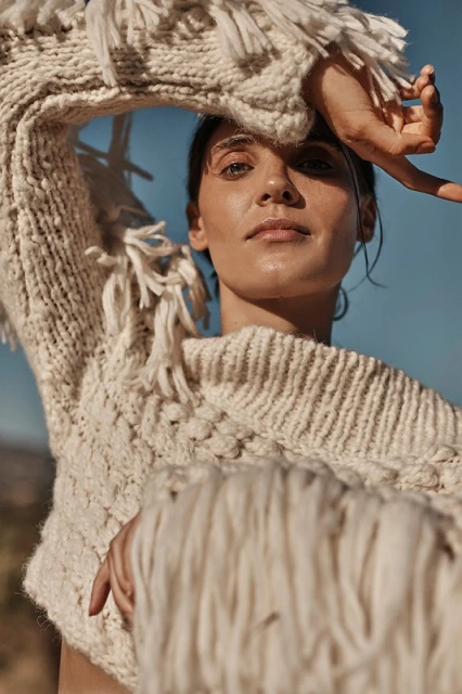 Woman in fringed knit sweater poses outdoors with blue sky. Sweater has chunky knit texture. Fashion, beauty, and natural light.