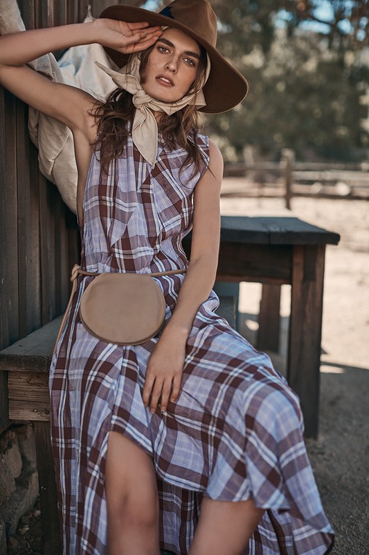 A fashionable adult female in a plaid dress, a belt bag, and a hat poses against a wood wall in an outdoor environment during daylight.