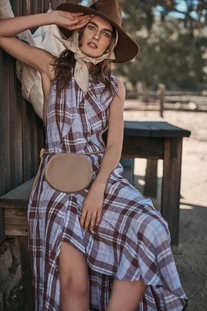 A fashionable adult female in a plaid dress, a belt bag, and a hat poses against a wood wall in an outdoor environment during daylight.