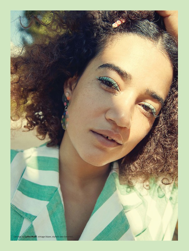 Close-up portrait of a woman with curly hair and green eye makeup, wearing a striped green and white shirt in an outdoor setting.
