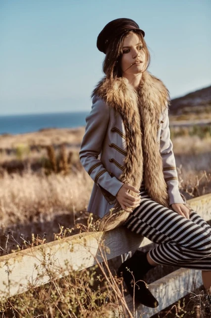 A woman poses outdoors, wearing a fur-collared jacket, black-and-white striped pants, and a cap, with a blurred background of the sea.