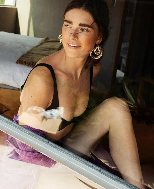 Adult woman in a black bikini top and wrap earrings poses inside a house with a white mattress. Sunlight highlights her face.