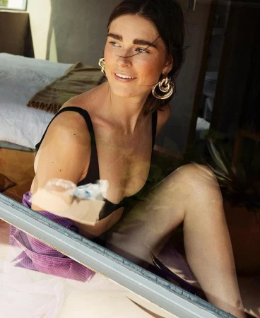 Adult woman in a black bikini top and wrap earrings poses inside a house with a white mattress. Sunlight highlights her face.