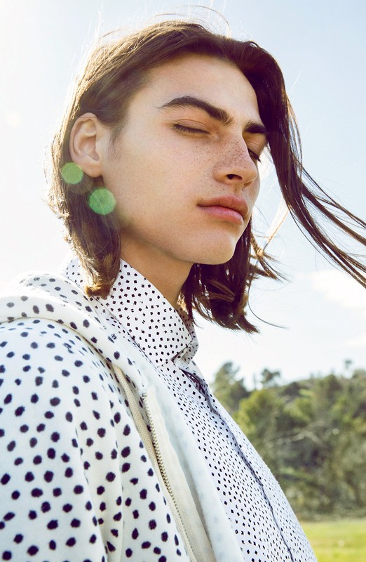 Portrait of a young adult male with long brown hair and freckles, wearing a polka dot shirt and jacket in an outdoor setting under bright sunlight.