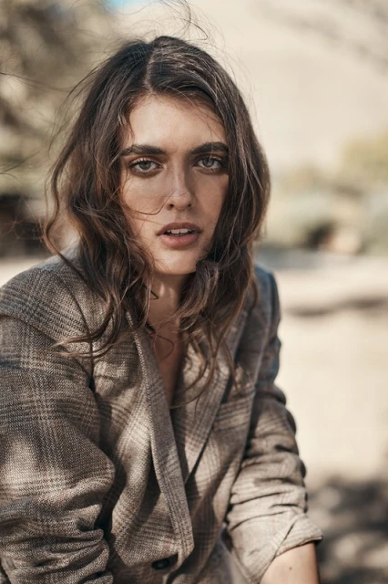 Portrait of a woman with wavy brunette hair in a brown plaid blazer with an open neckline, posing in an outdoor setting.