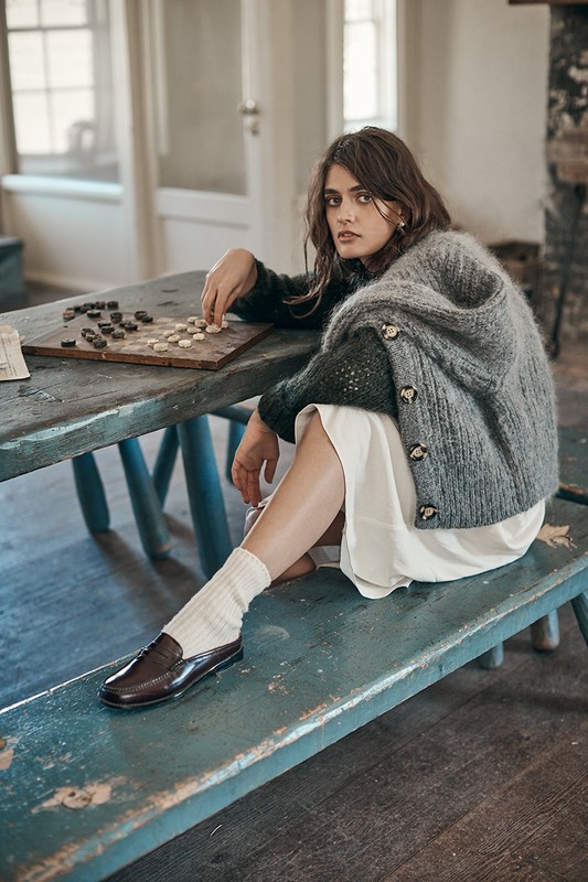 Woman in a cardigan sweater sits indoors at a table playing checkers, wearing a white dress, brown loafers, and white socks.