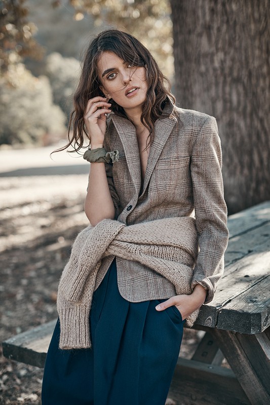 A woman in a blazer and wide-leg pants is sitting on a picnic table outdoors. She has a sweater tied around her shoulders.