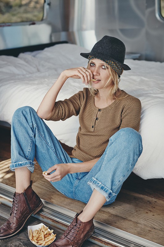 Blonde woman in hat and casual attire sits in an Airstream eating fries; wearing a neutral-tone shirt, blue jeans, and brown lace-up boots.