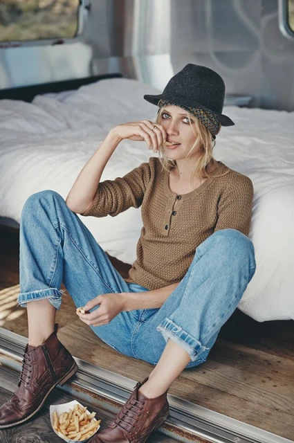 Blonde woman in hat and casual attire sits in an Airstream eating fries; wearing a neutral-tone shirt, blue jeans, and brown lace-up boots.