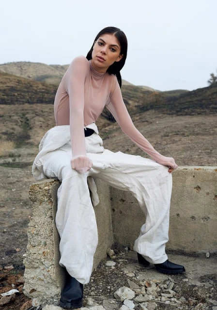A woman sits on a concrete structure wearing white overalls, a sheer top, and black boots in a barren, outdoor landscape with hills.