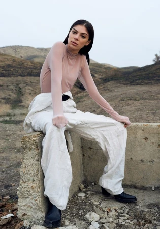 A woman sits on a concrete structure wearing white overalls, a sheer top, and black boots in a barren, outdoor landscape with hills.