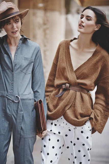 Two women pose outdoors in fashionable outfits: one in a denim jumpsuit and hat, the other in a brown sweater and polka dot pants.
