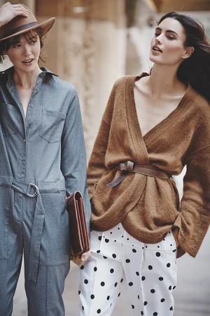 Two women pose outdoors in fashionable outfits: one in a denim jumpsuit and hat, the other in a brown sweater and polka dot pants.