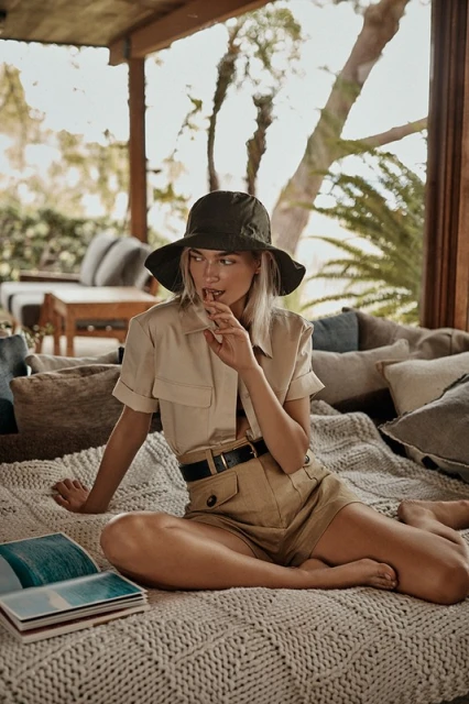 Adult female in a hat and casual summer outfit sits on a blanket outdoors with an open book in a relaxed setting.
