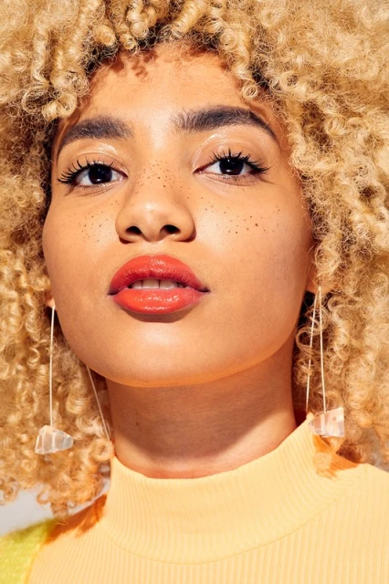 Close-up portrait of a young woman with curly blonde hair, wearing statement earrings and lipstick, in a brightly lit indoor setting.
