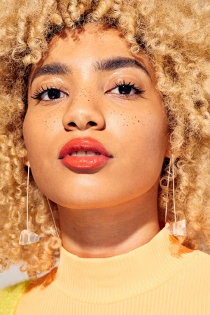 Close-up portrait of a young woman with curly blonde hair, wearing statement earrings and lipstick, in a brightly lit indoor setting.