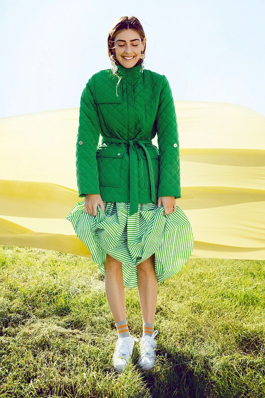 A woman in a green quilted jacket and striped skirt smiles in a grassy field with a yellow backdrop, creating a vibrant outdoor fashion portrait.