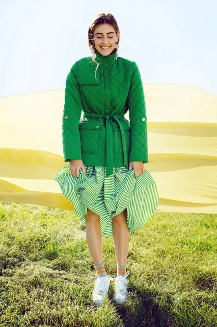 A woman in a green quilted jacket and striped skirt smiles in a grassy field with a yellow backdrop, creating a vibrant outdoor fashion portrait.