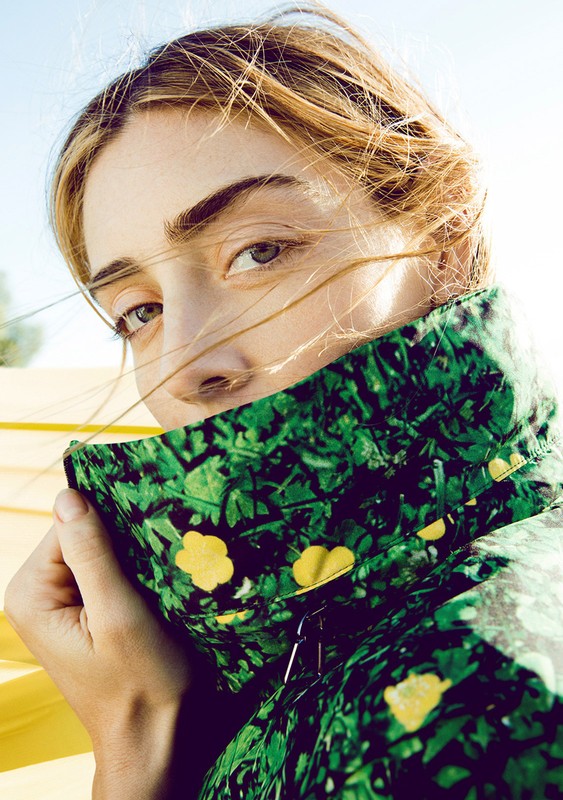 Close-up portrait of a blonde woman wearing a green floral jacket, posing outdoors with sunlight and a clear blue sky in the background.