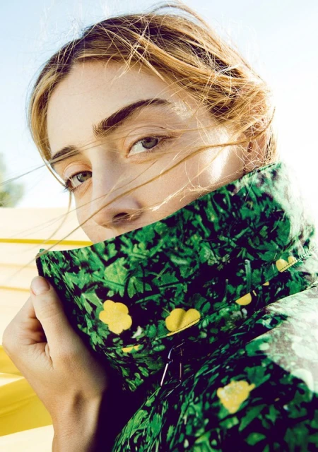 Close-up portrait of a blonde woman wearing a green floral jacket, posing outdoors with sunlight and a clear blue sky in the background.