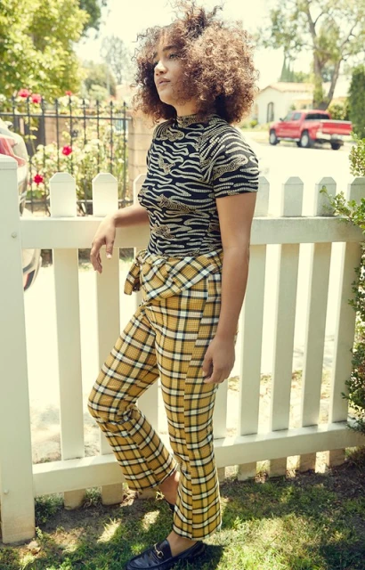Young woman with curly hair, wearing patterned top and plaid pants, stands in front of a white picket fence in a residential area.