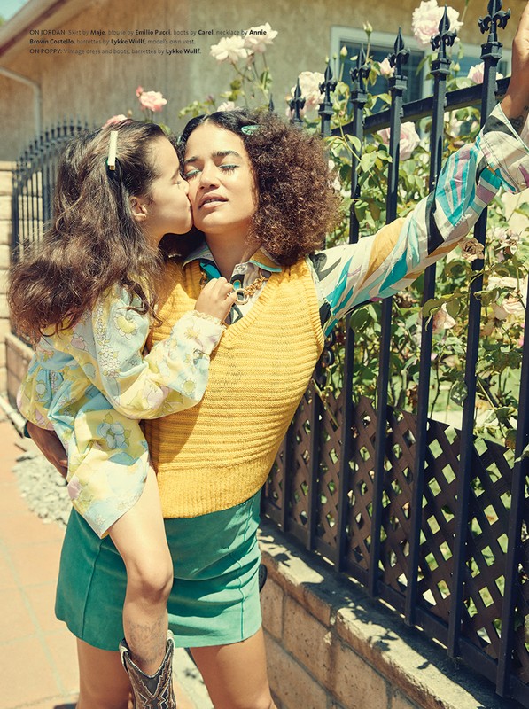 Mother in yellow sweater holds her daughter while she kisses her cheek outdoors near a black fence and white roses.