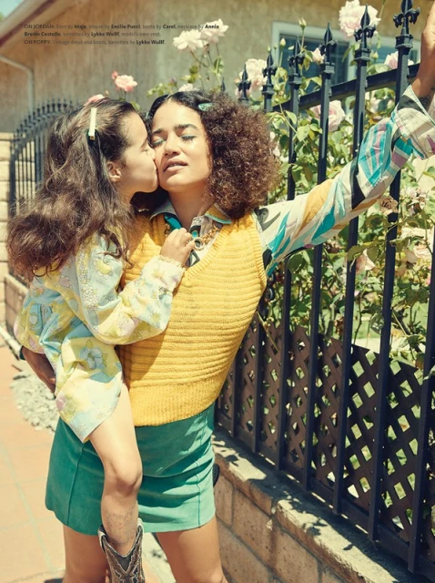 Mother in yellow sweater holds her daughter while she kisses her cheek outdoors near a black fence and white roses.